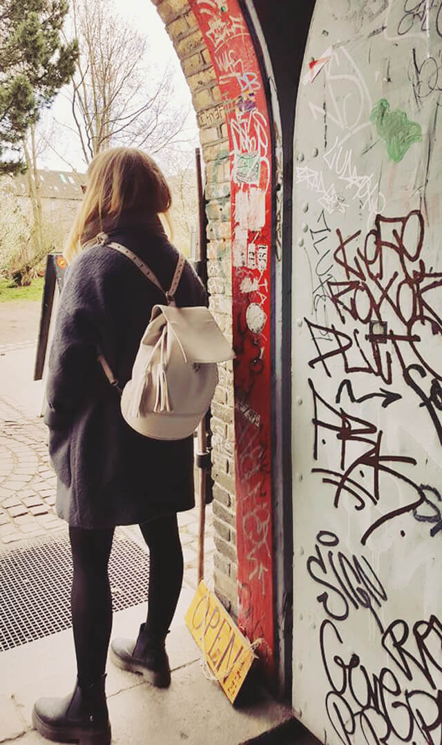 Lyzi stands looking away from camera in a doorway with graffiti on walls, she's wearing a big grey coat and a white leather backpack