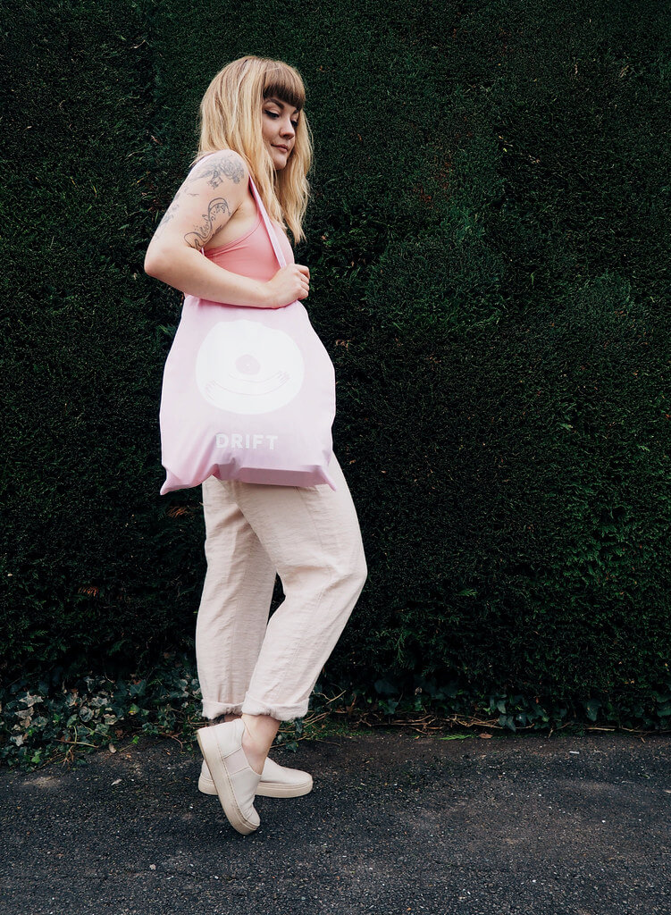 Lyzi wears a pink sports bra, neutral trousers and shoes, and pink Drift tote bag