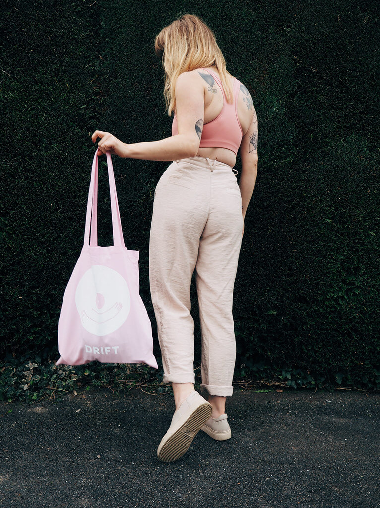 Lyzi wears a pink sports bra, neutral trousers and shoes, and pink Drift tote bag