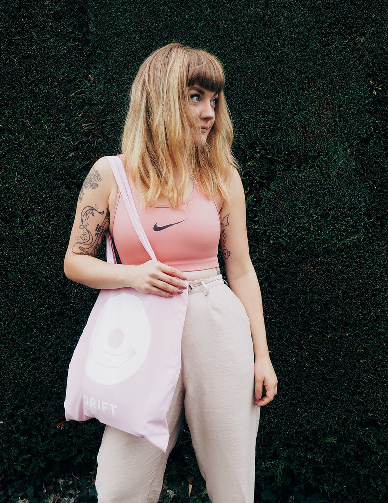 Lyzi wears a pink sports bra, neutral trousers and shoes, and pink Drift tote bag