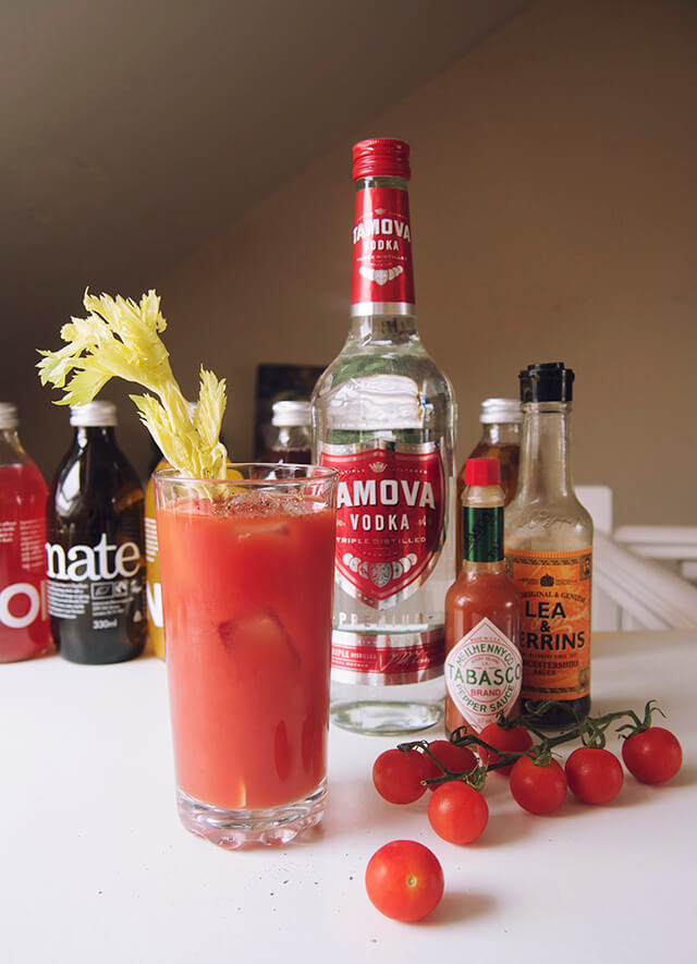 Ingredients next to a glass of a Bloody Mary - vodka, tabasco, tomatoes