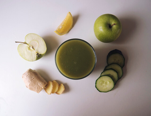 Green juice flatlay with glass of juice in the centre, surrounded by apple, ginger, lemon anc cucumber