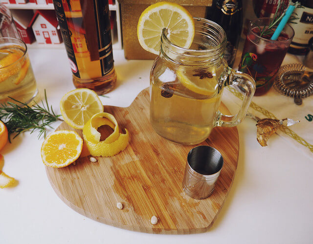 Cocktail making station, with heart-shaped wooden chopping board with sliced lemon, spirit measurer and mason glass with Warm Spice cocktail and a bottle of whisky