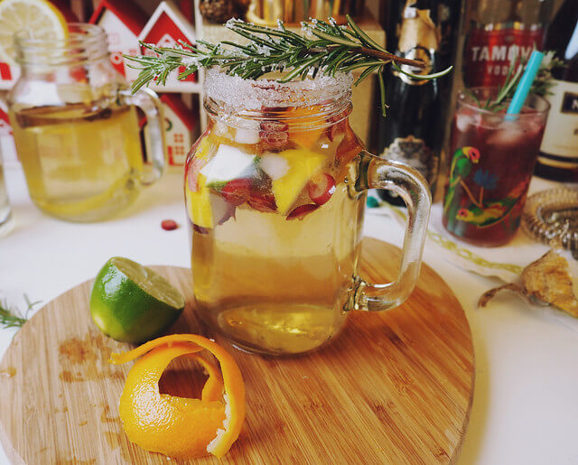 Cocktail making station, with wooden chopping board with sliced lime and orange, a mason glass with White Christmas Sangria cocktail garnished with rosemary, and more alcohol in the background