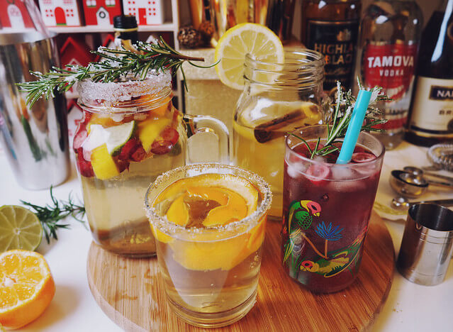 Four colourful Christmas cocktails on a wooden board with Christmas decorations and bottles of booze in the background