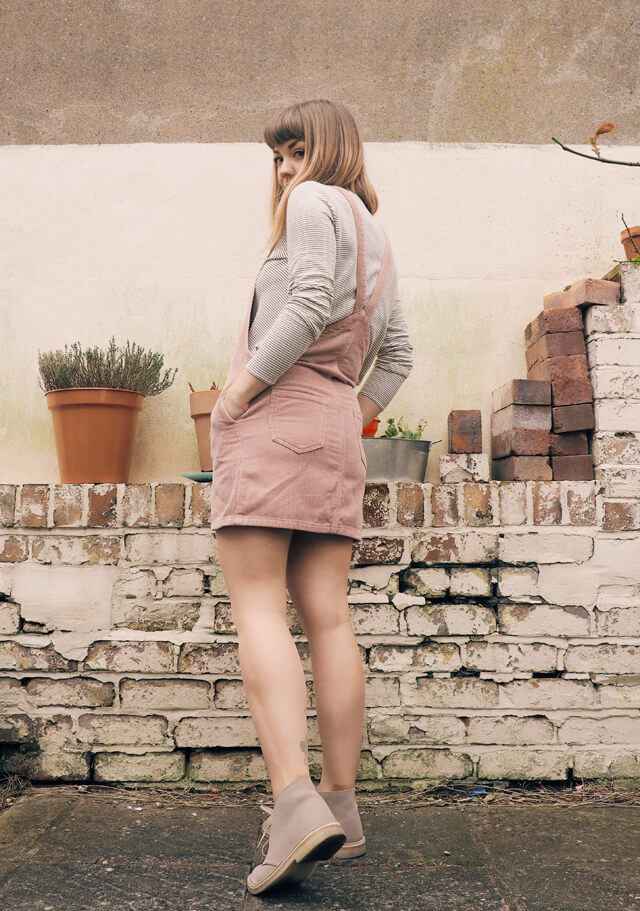 Back view of Lyzi in the garden wearing white top with black stripes underneath a short pink corduroy pinafore dress, and sand-coloured Clarks Desert Boots