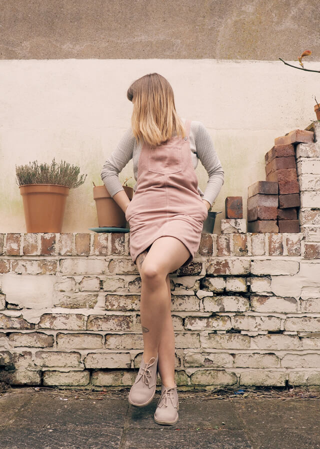 Front view of Lyzi in the garden wearing white top with black stripes underneath a short pink corduroy pinafore dress, and sand-coloured Clarks Desert Boots