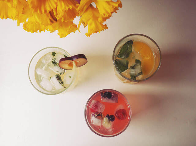 3 glasses of colourful fruity cocktails from above, with yellow daffodils creeping into shot