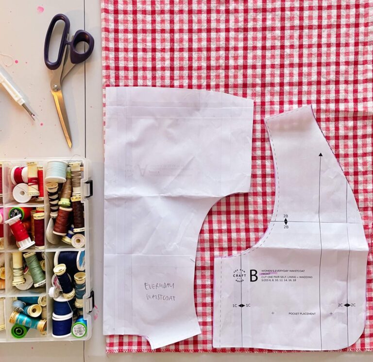 Everyday Waistcoat sewing pattern laid on red gingham, with threads and fabric scissors