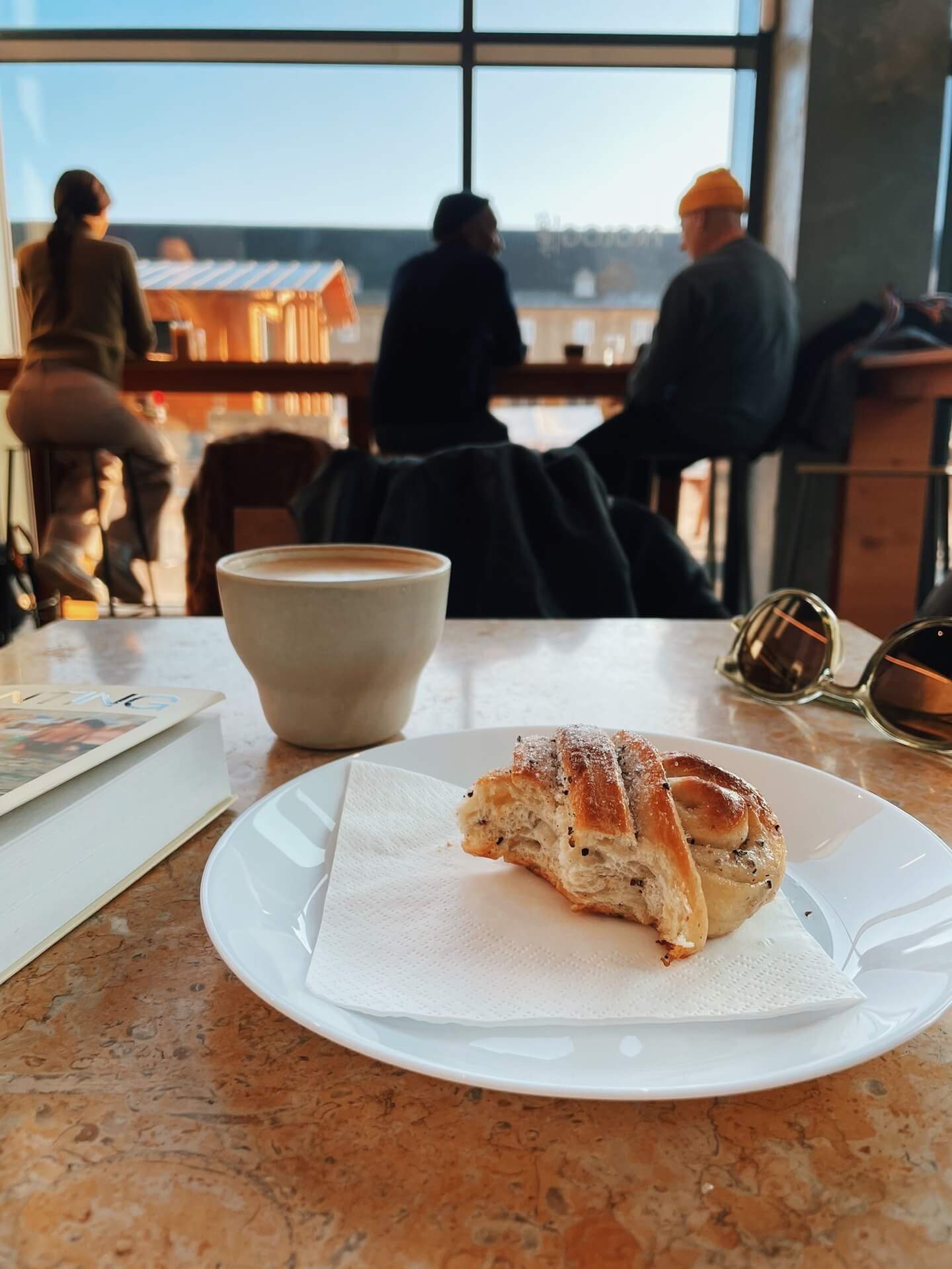 Prolog cafe scene with coffee and half-eaten cardamom bun in the foreground, and people sitting in the window in the background, out of focus